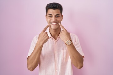 Young hispanic man standing over pink background smiling with open mouth, fingers pointing and forcing cheerful smile