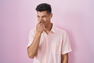Young hispanic man standing over pink background smelling something stinky and disgusting,...