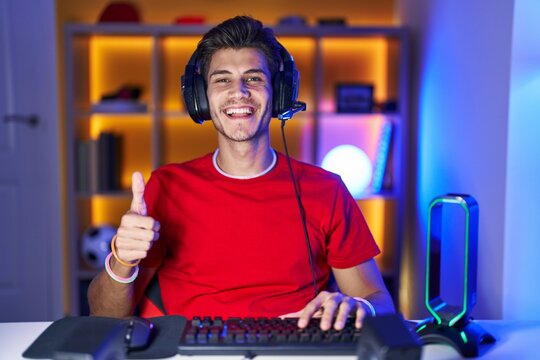 Young hispanic man playing video games doing happy thumbs up gesture with hand. approving expression looking at the camera showing success. - Powered by Adobe