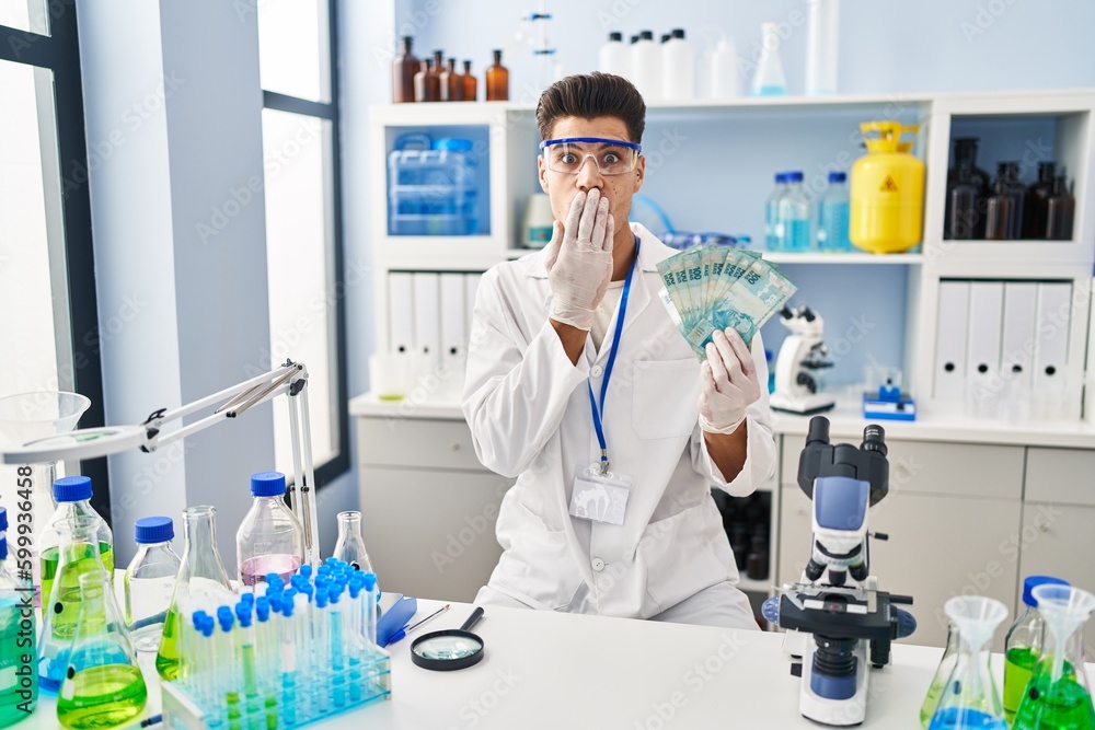 Canvas Prints Young hispanic man working at scientist laboratory holding brazilian reals covering mouth with hand, shocked and afraid for mistake. surprised expression