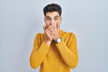 Young hispanic man standing over blue background shocked covering mouth with hands for mistake....