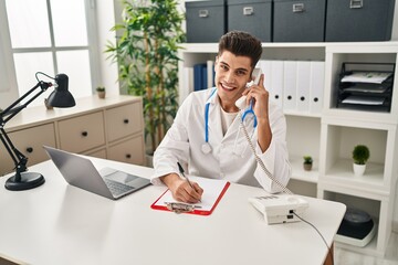 Young hispanic man wearing doctor uniform talking on the telephone at clinic