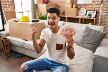 Young hispanic man moving to a new home holding shekels pointing thumb up to the side smiling happy with open mouth