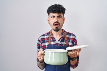 Young hispanic man with beard wearing apron holding cooking pot in shock face, looking skeptical...