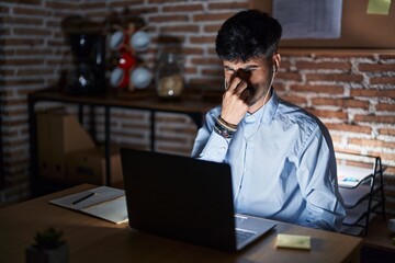 Young hispanic man with beard working at the office at night smelling something stinky and disgusting, intolerable smell, holding breath with fingers on nose. bad smell