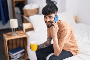 Young hispanic man talking on smartphone drinking coffee at bedroom