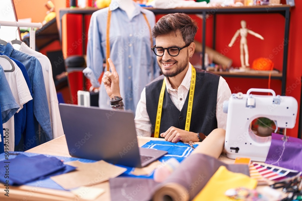 Sticker Young hispanic man with tattoos dressmaker designer using laptop at tailor room smiling happy pointing with hand and finger to the side