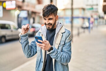 Young hispanic man using smartphone with winner expression at street