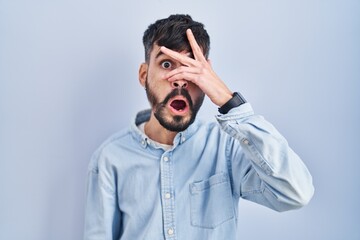 Young hispanic man with beard standing over blue background peeking in shock covering face and eyes...
