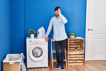 Young hispanic man with beard doing laundry standing at laundry room smiling happy doing ok sign with hand on eye looking through fingers