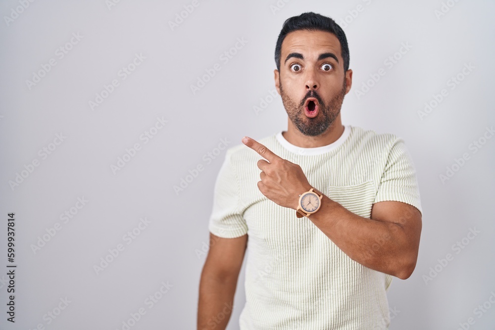 Canvas Prints Hispanic man with beard standing over isolated background surprised pointing with finger to the side, open mouth amazed expression.