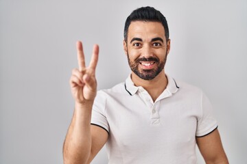Fototapeta premium Young hispanic man with beard wearing casual clothes over white background showing and pointing up with fingers number two while smiling confident and happy.