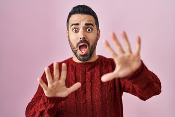 Young hispanic man with beard wearing casual sweater over pink background afraid and terrified with fear expression stop gesture with hands, shouting in shock. panic concept.