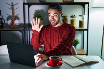 Young hispanic man with beard using computer laptop at night at home waiving saying hello happy and smiling, friendly welcome gesture
