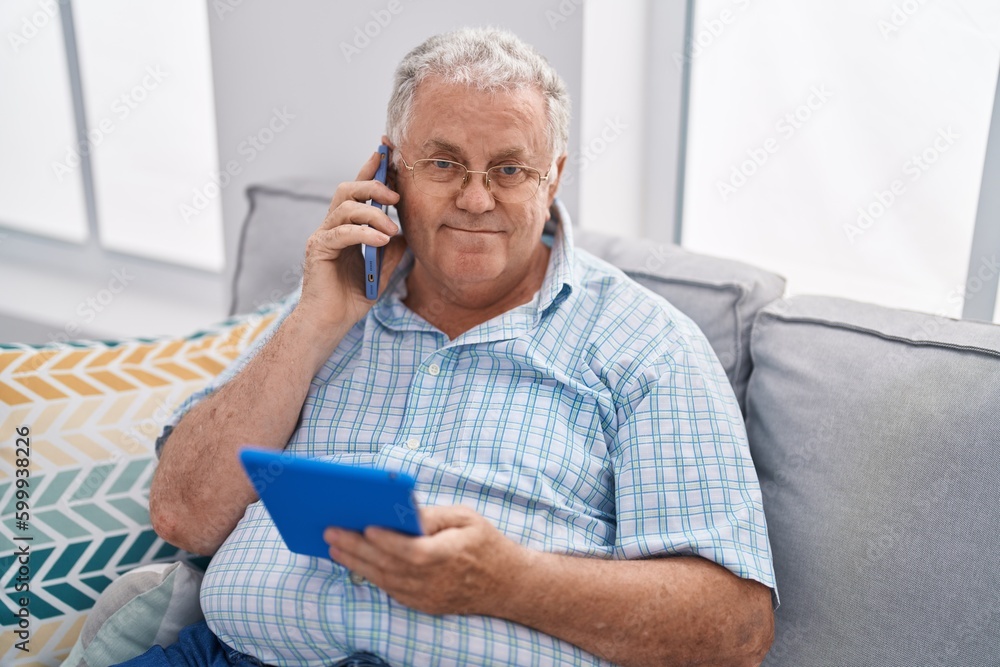 Poster middle age grey-haired man talking on smartphone using touchpad at home