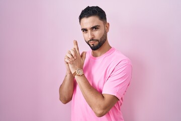 Hispanic young man standing over pink background holding symbolic gun with hand gesture, playing killing shooting weapons, angry face