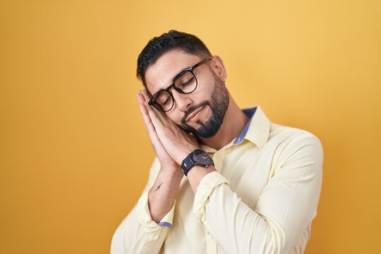 Hispanic young man wearing business clothes and glasses sleeping tired dreaming and posing with hands together while smiling with closed eyes.