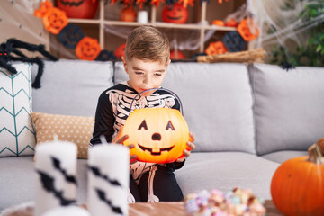 Fototapeta premium Adorable hispanic boy wearing skeleton costume looking pumpkin basket with surprise expression at home