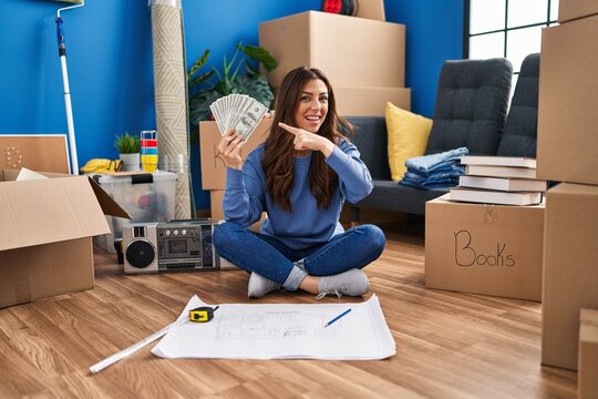 Young brunette woman sitting on the floor at new home holding money smiling happy pointing with hand and finger