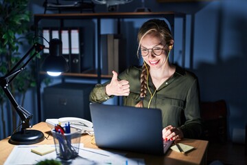 Young blonde woman working at the office at night doing happy thumbs up gesture with hand. approving expression looking at the camera showing success.
