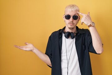Young caucasian man wearing sunglasses standing over yellow background confused and annoyed with open palm showing copy space and pointing finger to forehead. think about it.
