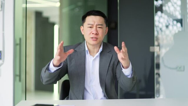 Webcam view. Angry asian man in formal suit arguing talking on video call while sitting at workplace in modern office. Excited disgruntled businessman, owner, boss quarreling, expressing a problem