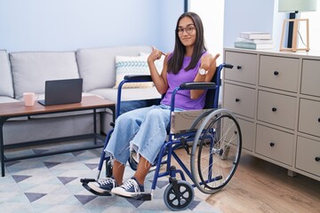 Young hispanic woman sitting on wheelchair at home looking confident with smile on face, pointing...