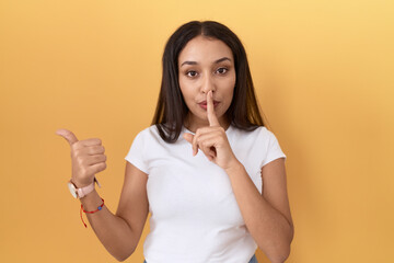 Young arab woman wearing casual white t shirt over yellow background asking to be quiet with finger on lips pointing with hand to the side. silence and secret concept.