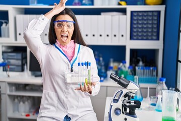 Young brunette woman working at scientist laboratory stressed and frustrated with hand on head, surprised and angry face