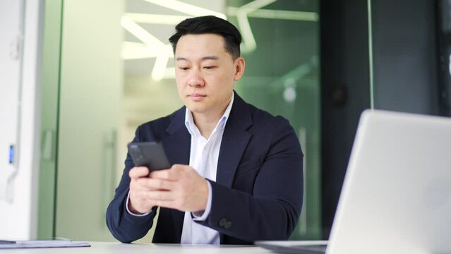 Serious asian businessman in a formal suit is using a browsing smartphone while sitting at a workplace in a office. Concentrated pensive owner reading, checking email or typing text message to client