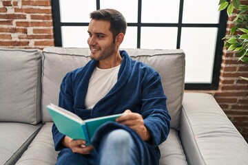 Young hispanic man wearing bathrobe reading book at home