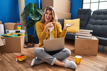 Young woman sitting on the floor at new home using laptop amazed and smiling to the camera while presenting with hand and pointing with finger.