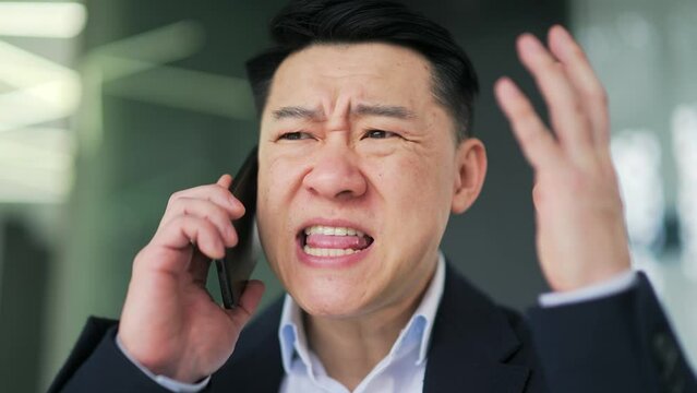 Close up. Angry asian man in formal suit arguing while talking on smartphone in modern office. Excited, dissatisfied businessman, owner, boss shouts into the phone, quarrels, expresses a problem