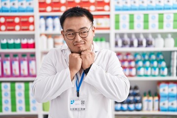 Chinese young man working at pharmacy drugstore laughing nervous and excited with hands on chin looking to the side