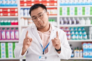Fototapeta premium Chinese young man working at pharmacy drugstore pointing up looking sad and upset, indicating direction with fingers, unhappy and depressed.