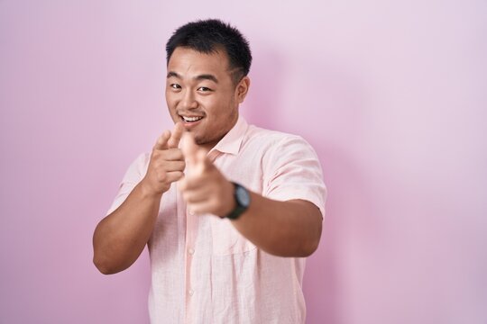 Chinese young man standing over pink background pointing fingers to camera with happy and funny face. good energy and vibes.