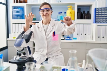 Hispanic girl with down syndrome working at scientist laboratory showing and pointing up with fingers number six while smiling confident and happy.