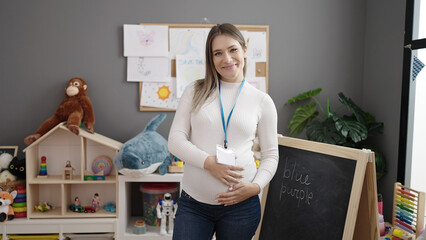 Young pregnant woman preschool teacher smiling confident touching belly at kindergarten