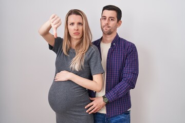 Young couple expecting a baby standing over white background looking unhappy and angry showing rejection and negative with thumbs down gesture. bad expression.