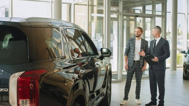 Successful middle-aged businessman in a business suit is selling cars to a young hansome male buyer in a car showroom. Business man chosing a new car standing with salesperson in the car.