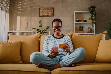 Obraz premium African american woman using credit card and asmartphone while online shopping at home
