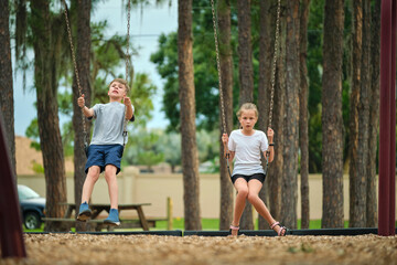 Fototapeta na wymiar Two young teenage children, girl and boy playing on swings together outdoors on bright sunny vacations day