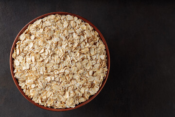 Oat flakes in a clay bowl on a dark background. Organic rolled oats for healthy nutrition. Copy space. Top view