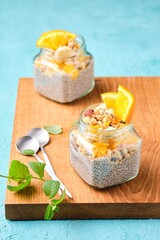 Obraz na płótnie Canvas Healthy food, chia pudding on almond milk with orange, banana and granola in glass jars on a wooden board on a turquoise concrete background.