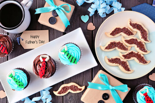 Fathers Day dessert table scene. Top view on a dark wood background. Gifts, greeting card, shirt cupcakes and mustache cookies.