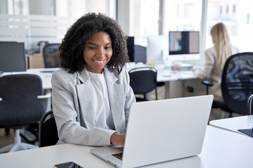 African American young female businesswoman, successful entrepreneur using app gadget sitting at work place in office. Middle age business woman working, browsing, and typing on touchscreen pc laptop.