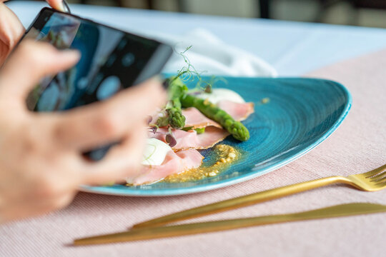 Appetizer with rosemary ham, asparagus, peanuts, and taleggio on the blue plate. Pink cloth with golden cutlery. A woman taking a photo of the food with a mobile phone.