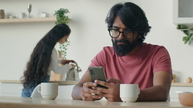 Bearded muslim Indian Arabian man remote working from home kitchen use mobile phone apps cellphone chatting online. Ethnic woman wife housewife cooking morning coffee breakfast for husband background