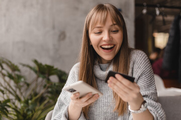 Happy young blonde woman with bang customer shopper holding credit card using cell phone mobile app buying fashion clothes paying online make purchase in ecommerce digital store on smartphone in cafe.