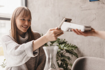 Blonde attractive woman paying with cellphone resting in cafe while waitress holding payment terminal. Girl relaxing in restaurant. Focus on part payment terminal and mobile phone.